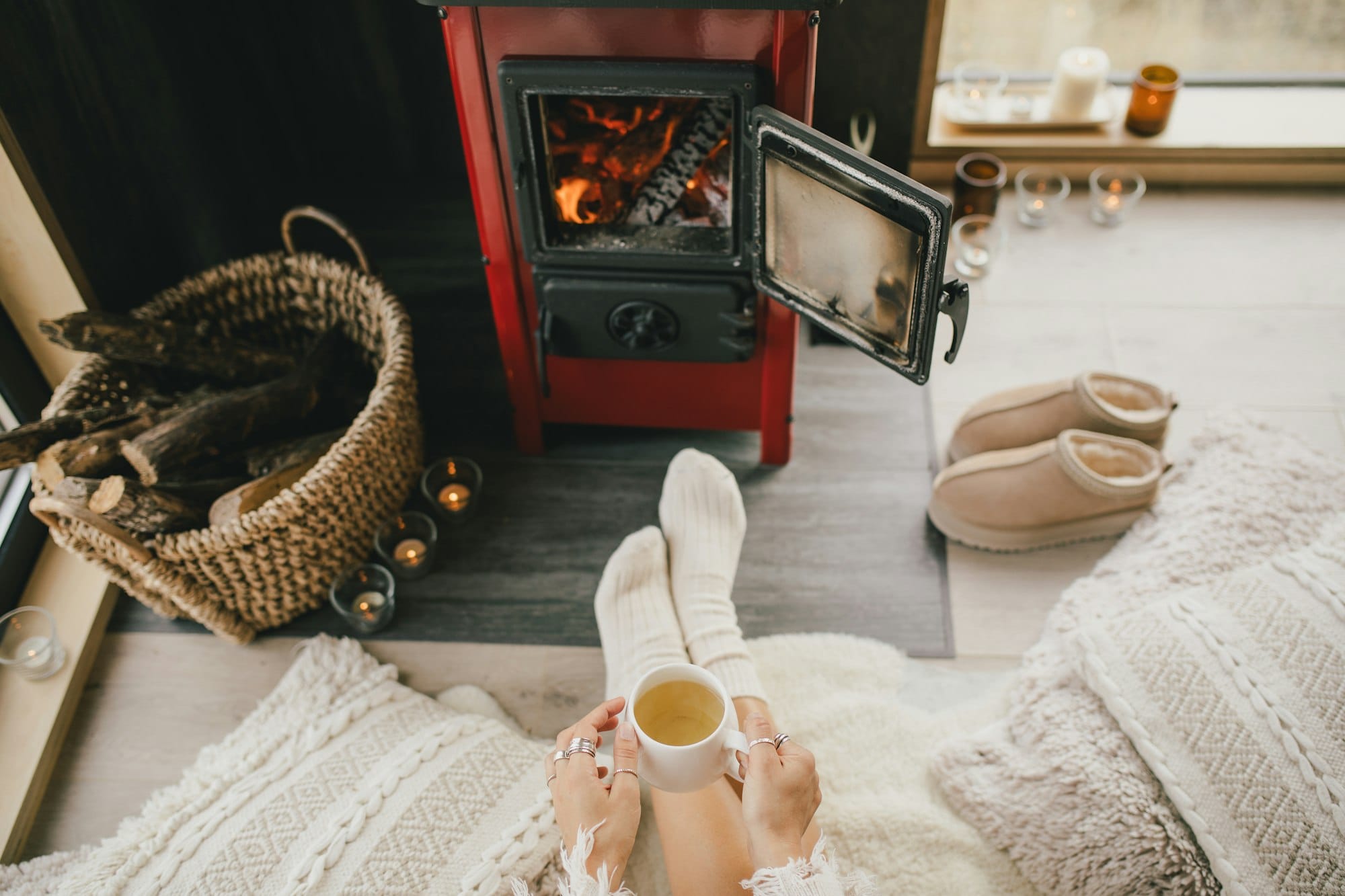 Young woman sitting by the fireplace in white sweater, drinking tea in cozy log cabin.